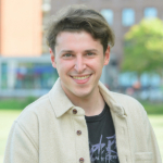 A colour portrait of Ellis Karran with trees and buildings in the background. Ellis is a white man with brown hair who is wearing a cream jacket over a dark t-shirt.
