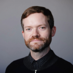 A colour portrait on a grey background of Ian Chamberlain. Ian a white man with red hair and beard, wearing a dark jumper