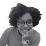 A young black woman smiles at the camera with her chin resting on her first.  She has dark hairm glasses and a white watch on and is wearing a light jumper