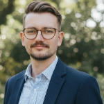 A colour photograph of James Sharp with trees in the background. James is a white man with brown hair, beard and glasses who is wearing a navy blazer over a blue check shirt.
