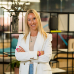A colour portrait of Jane Hunt stood smiling and with her arms folded in an open-plan office. Jane is a white woman with long blond hair who is wearing a white jacket.