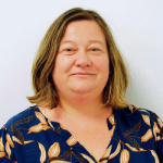 A colour portrait of Jenny Manchester. Jenny is a white woman with shoulder length hair who is wearing a blue and orange patterned blouse.