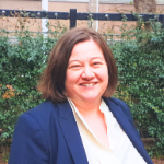 A colour portrait of Jenny Manchester stood in front of a hedge. Jenny is a white woman with brown bobbed hair who wears a blue jacket over a white top.