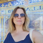A portrait of Kat Dibbits. Kat is a white woman with blonde hair, wearing sunglasses and stood in front of blue and yellow tiles