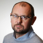 A colour portrait of Marc Evans on a grey background. Marc is a white man with receding dark hair who wears glasses and a grey jumper over a blue collared shirt.