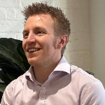 A colour portrait of Max Jewell photographed from the right side of his face. Max is a white man with blond hair who is wearing a pink shirt.