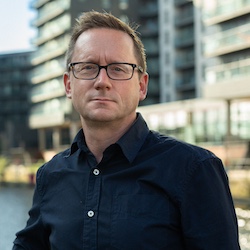 A colour portrait of Nev Ridley. Nev is a white man with short blond hair who wears glasses and a dark blue shirt. He is stood in front of modern dockside buildings