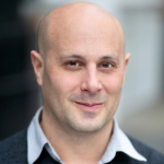 A colour portrait of Nick Gold. Nick is a white man with wearing a light blue shirt and dark top