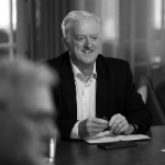 A black and white portrait of Nigel Sarbutts sat at a table. Nigel is a white man with light hair who is wearing a dark jacket and white shirt.