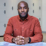 A colour portrait of Richard Etienne sat at a table looking at the camera. Richard is a Black man wearing an orange shirt. The background is white with dark flecks.