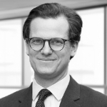 A black and white photograph of Ryan McSharry. Ryan is a white man with short dark hair, wearing glasses, a dark suit jacket and tie.