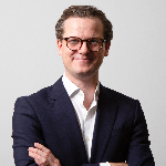 A colour portrait against a grey background of Ryan McSharry stood with his arms crossed. Ryan is a white man with short hair who is wearing glasses and a dark jacket.