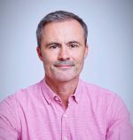 A portrait of Ben Verinder on a grey background. Ben is a white man with grey hair and stubble wearing a pink shirt