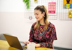 Side angle of Beth Turner, a white woman with long brown hair and wearing a mauve and blue patterned dress, sits at a laptop. There is a mug on the table and pictures on the white wall behind her