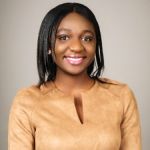 A portrait of a smiling Esenam Agubretu against a grey background. Esenam is a Black woman with shoulder length dark hair and wearing a gold top