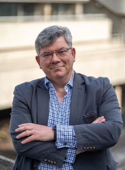 Guy Clapperton, a middle aged white man with grey hair and glasses, stood with his arms crossed across his chest. Guy wears a grey suit and blue check shirt