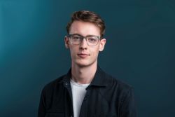 Joel Meekison is a young white man with short light brown hair. He is wearing glasses, a white t-shirt and a black shirt, he is against a blue background