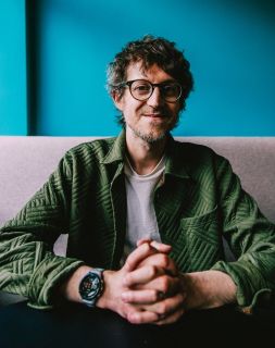 A portrait of Leigh Greenwood, a white man with glasses and dark curly hair, wearing a green jacket. His hands are clasped in front of him. He is sat on a grey sofa with a dark turquoise wall behind him 