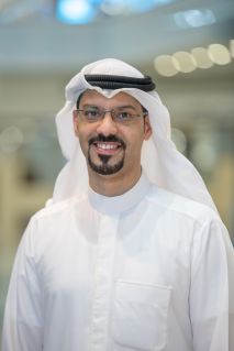A portrait of Saad smiling at the camera. Saad is a Kuwaiti man with glasses and dark beard who wears a white ghutrah or headscarf secured with a black agal chord. He wears a white kandura buttoned jacket