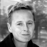 A black and white portrait of Christian Koch. Christian is a white man with short hair. There are trees in the background