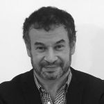 A black and white portrait of Ryan Herman. Ryan is a white man with short dark hair who wears a dark jacket over a check shirt