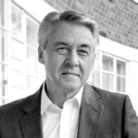 A black and white photograph of Jon White, a white middle aged man with grey hair, white shirt and dark blazer in the open air. Behind him are windows and bricks of a building