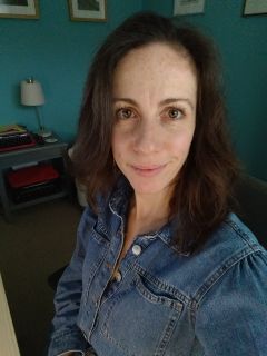Kate Hughes looking at the camera. She has brown hair and is wearing a denim shirt. The wall in the background is turquoise with four picture frames. There is a lamp and a table in the background.