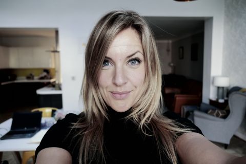 Rebecca Robers taking a selfie. She is a white woman with blonde hair and wears a short sleeve black top. A kitchen and living room are in the background