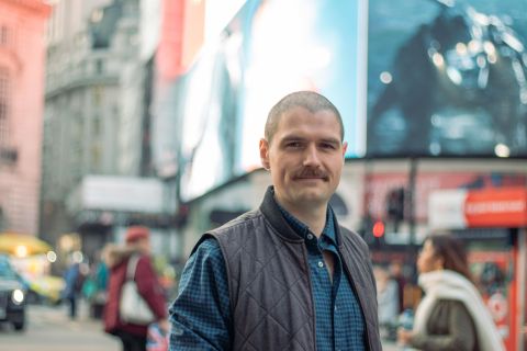 Sam Patchet stood in front of the screens at Piccadilly Circus. Sam is a white man with cropped dark hair and moustache. He wears a dark gilet over a blue shirt.