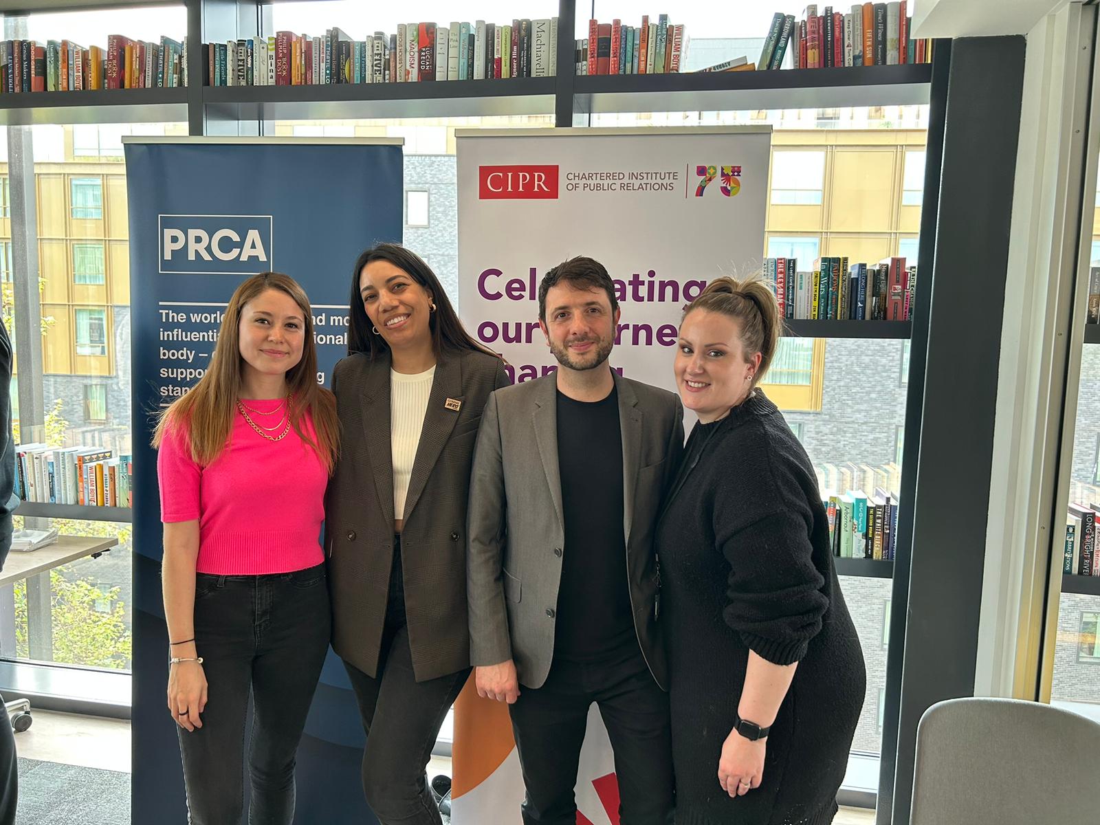 Four people stood smiling at the camera in front of a book case and signs from CIPR and PRCA