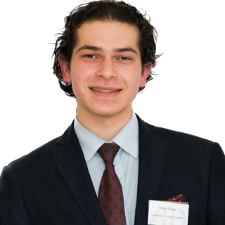 Yanis Fekar, a white man with dark hair and wearing a dark suit, blue shirt and red and black tie, smiles.