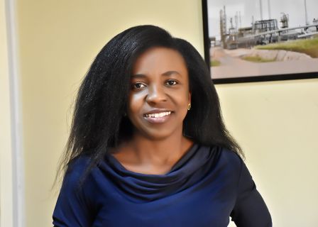 Matilda Adjoah, a Black woman with dark hair and wearing a navy top, looking directly at the camera and smiling. In the backround is a yellow wall with a picture frame on the right.