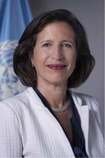 Melissa Fleming, a white woman with dark hair and wearing a white jacket, smiles while facing the camera. In the background is a blue and white UN flag.