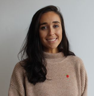 Head and shoulders photo of Ruby Kite looking straight at the camera. She has long dark hair and is wearing a roll-neck jumper with a poppy broach
