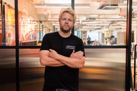 James Kirkham, a white man with blond hair, wears a black t-shirt with his arms folded. He is stood in front of a glass office partition.