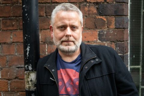 Richard Stone, a white man with grey hair and beard, wearing a jacket and t-shirt. He is stood in front of a brick wall.