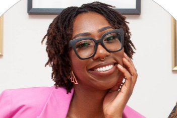 Subira Jones, a Black woman, wearing glasses and smiling. She wears a pink jacket. Her left hand is rested on the side of her face