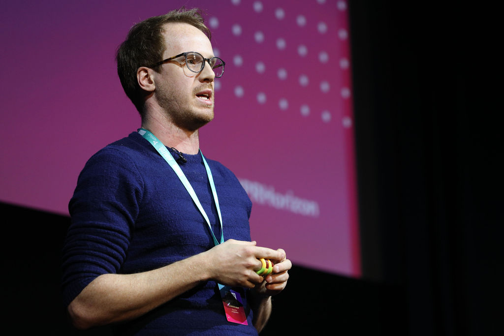Alex Partridge, a white man with brown hair, who wears glasses and a navy jumper. He holds a yellow fidget toy in his clasped hand. A large pink screen is in the background