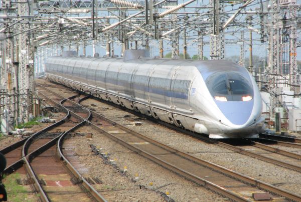 A streamlined Shinkansen bullet train runs under wires help up by metal gantries. There are five railway tracks and two sets of points in the foreground
