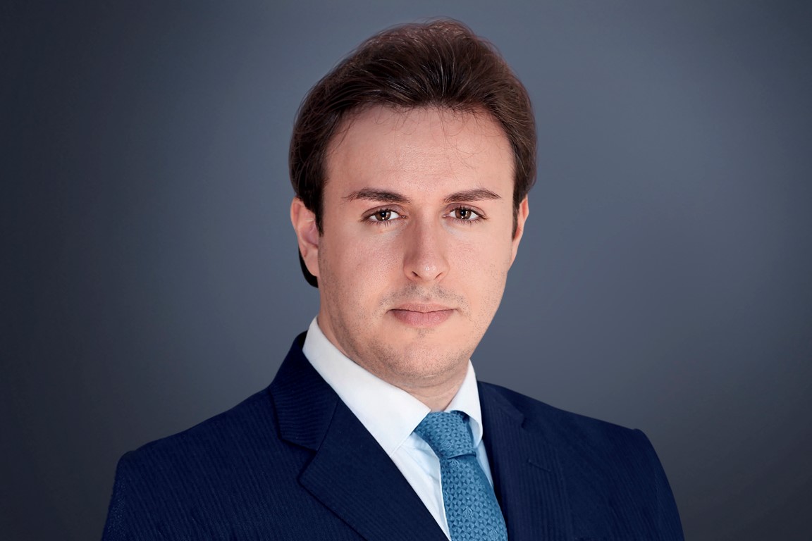 Alex Malouf, a white man with brown hair, against a dark grey background. Alex wears a navy suit and mid-blue tie