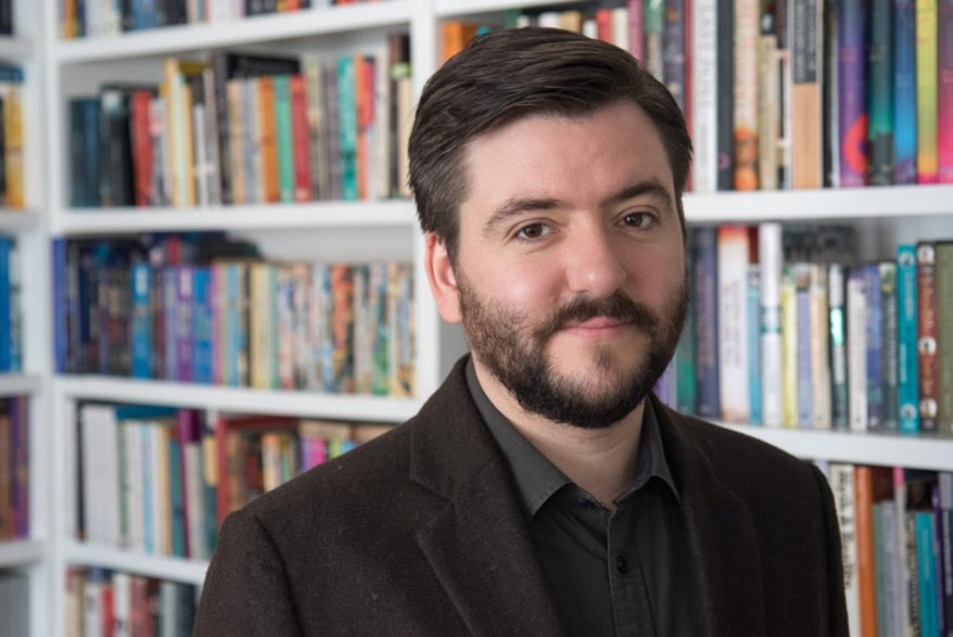 Andrew Copson in front of a full bookcase. Andrew is a white man with dark hair and beard. He wears a brown jacket and shirt