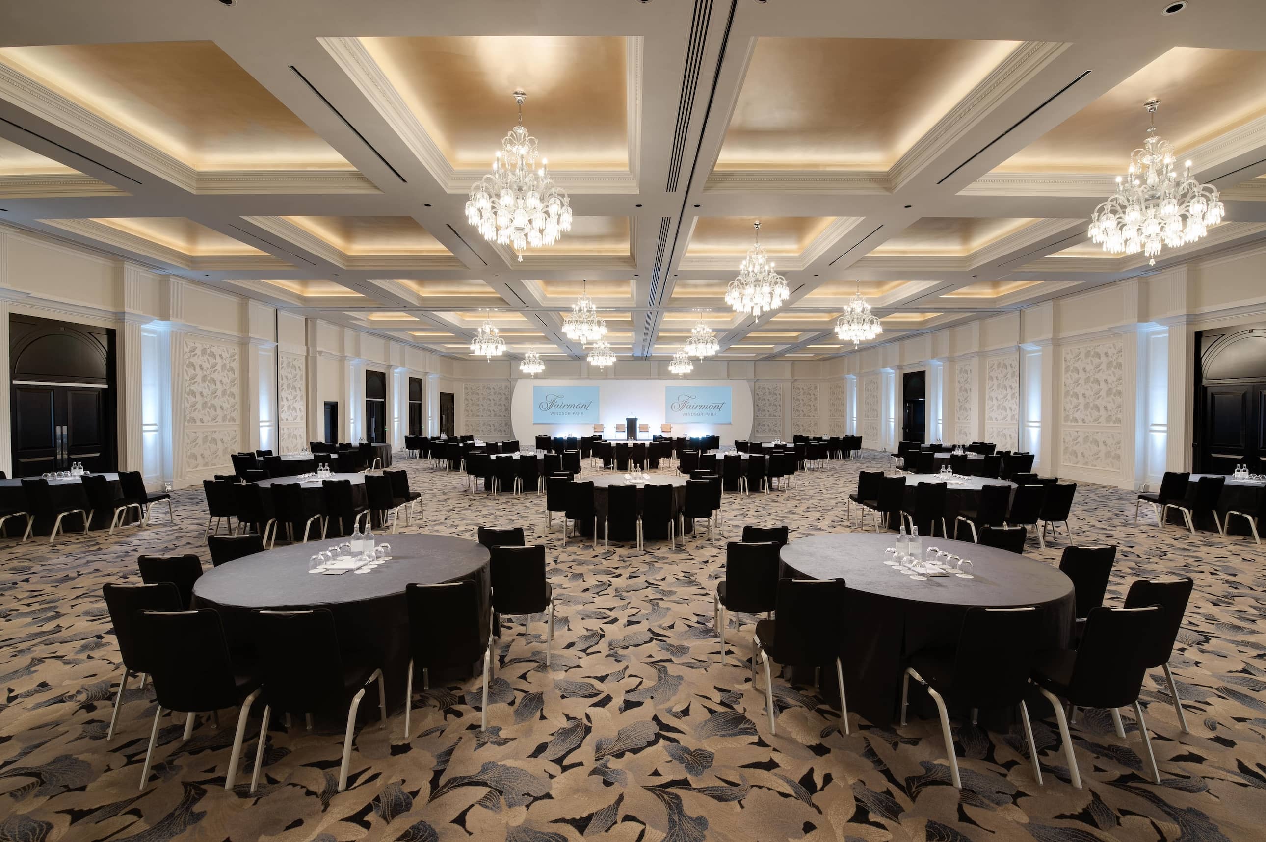 Large conference room with white and grey patterned carpet, more than a dozen large tables each with at least seven black chairs facing the front stage. The ceiling is panelled with 10 glass chandeliers