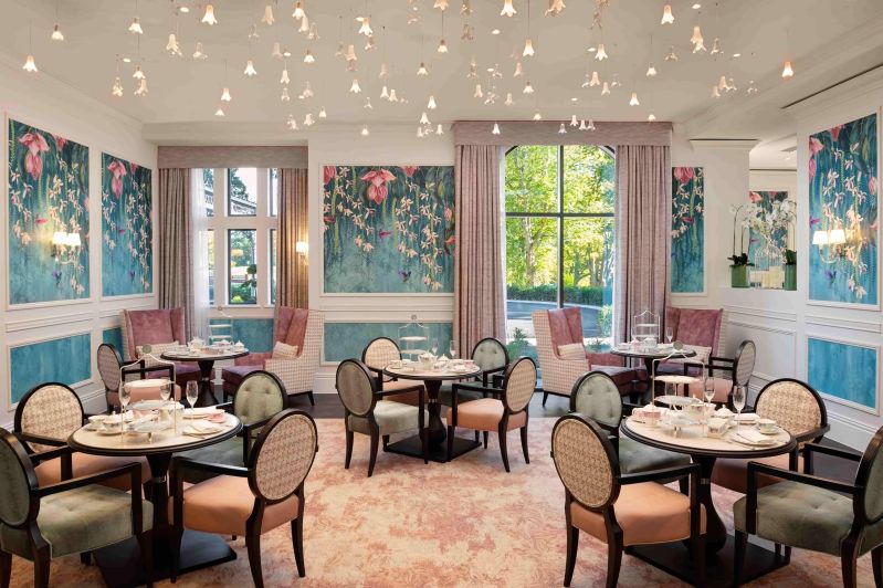 A dining room with two windows, drapes, dozens of small hanging white lights, three sets of four tables and chairs and two sets of armchairs with small dining tables. The pastel colour palette include blue, pink, orange and green heels with turquoise wall panelling decorated with pink orchids