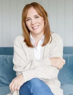 Natalie Chisolm, a smiling white woman with red shoulder length hair. She wears a cream cardigan and blue jeans while sat on a blue sofa