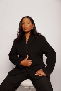 Sharmadean Reid sat on a white stool on a grey background. Reid is a Black woman, with long hair, who wears a black jacket and trousers.