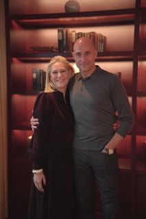 Tanya Rose, a white woman with blonde hair and dark suit, hugs Mark Strong, a white, clean shaven man, who wears a grey top. There is a bookcase behind them
