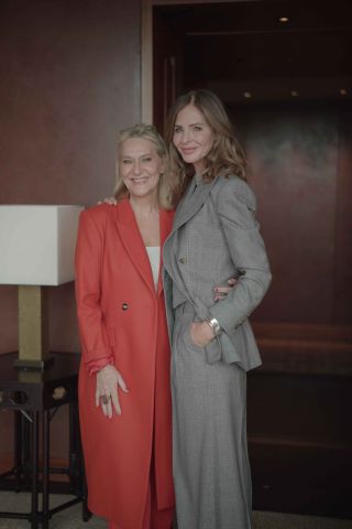 Trinny Hall, a white woman with blonde hair and wearing a red coat, smiles, while hugging Trinny Hall, a white woman with shoulder length brown hair, wearing a two-piece grey jacket and trouser suit