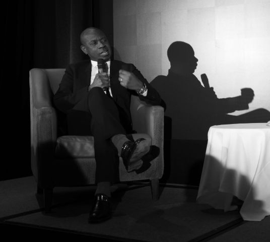 A black and white photo of Clive Myrie sat in an armchair holding a microphone. To his right is a perfect shadow of him.