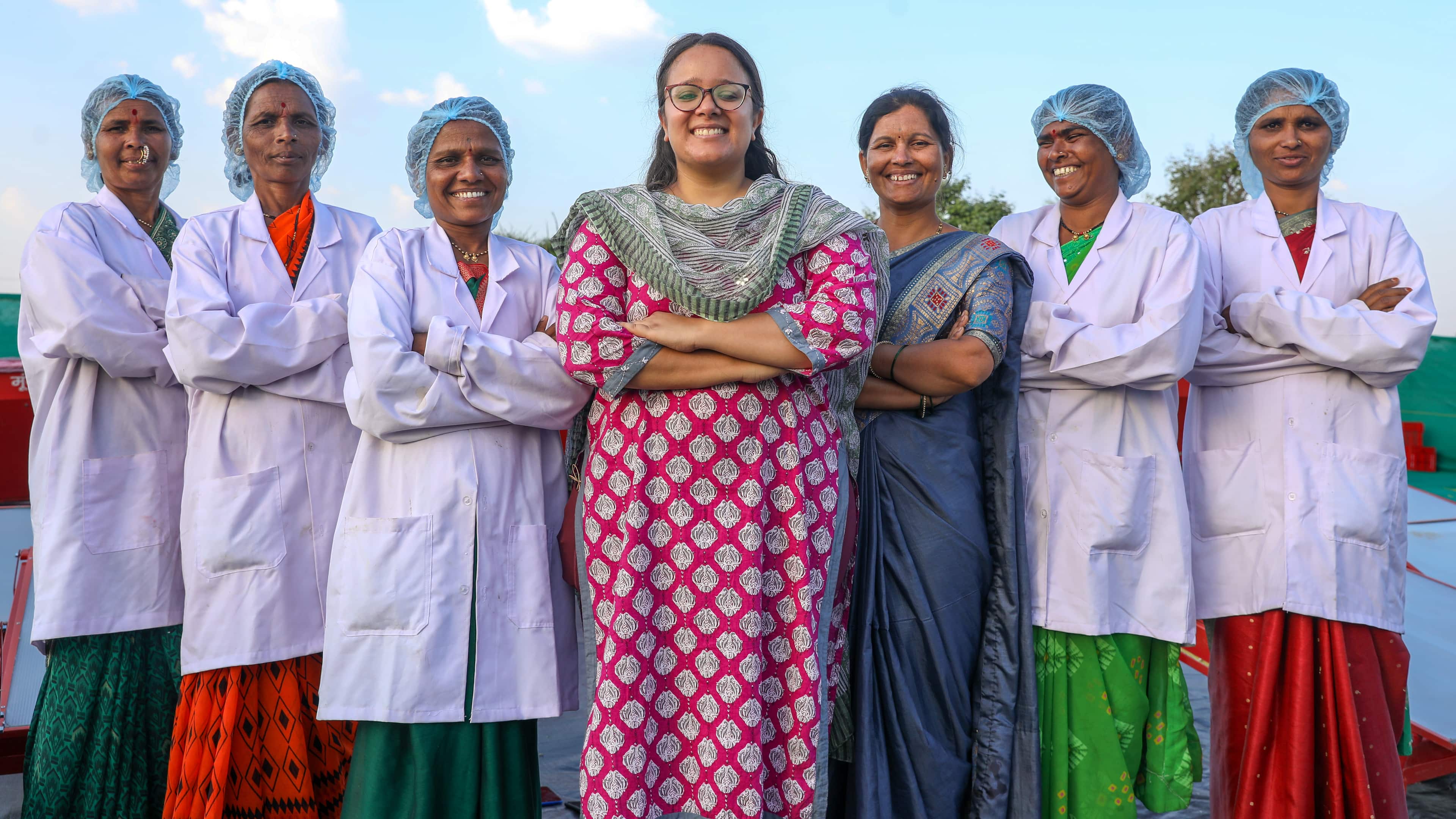 Seven Asian women smiling at the camera with Nidhi Plant in the centre. All wear bright coloured sari dresses. Three on the left and two on the right wear white jackets and blue hair nets.