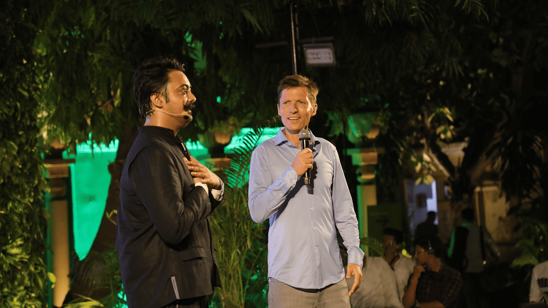 Tamseel Hussain, an Asian man with short hair and moustache, wears a black jacket, stood next to Colin Butfield, a white man with short brown hair, blue shirt and holding a microphone. They are on stage surrounded by plants, green light and an audience